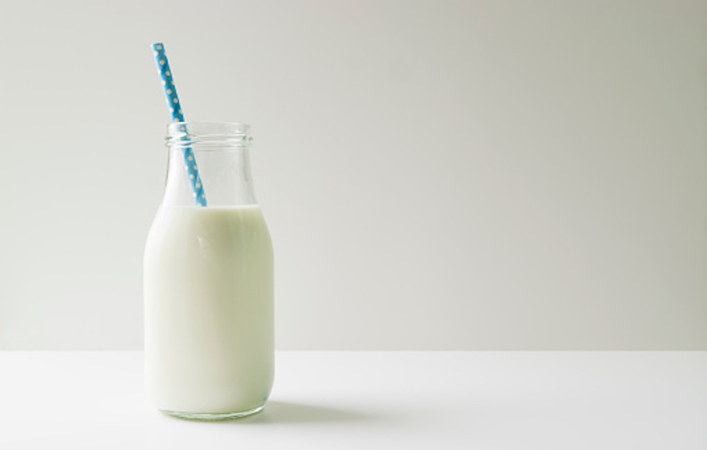 Clear, glass bottle of milk with a blue polka a dot straw set against a white backdrop