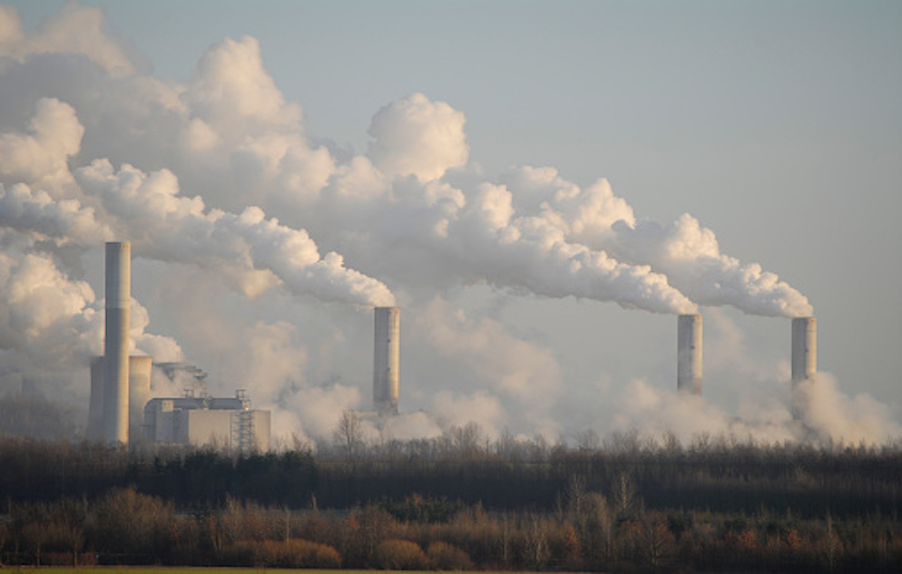 Five silos blowing air pollution over a tree-lined landscape.
