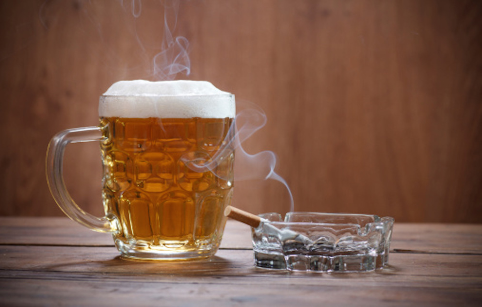 Clear mug of beer next to a smoking cigarette on a clear astray atop wooden surface.