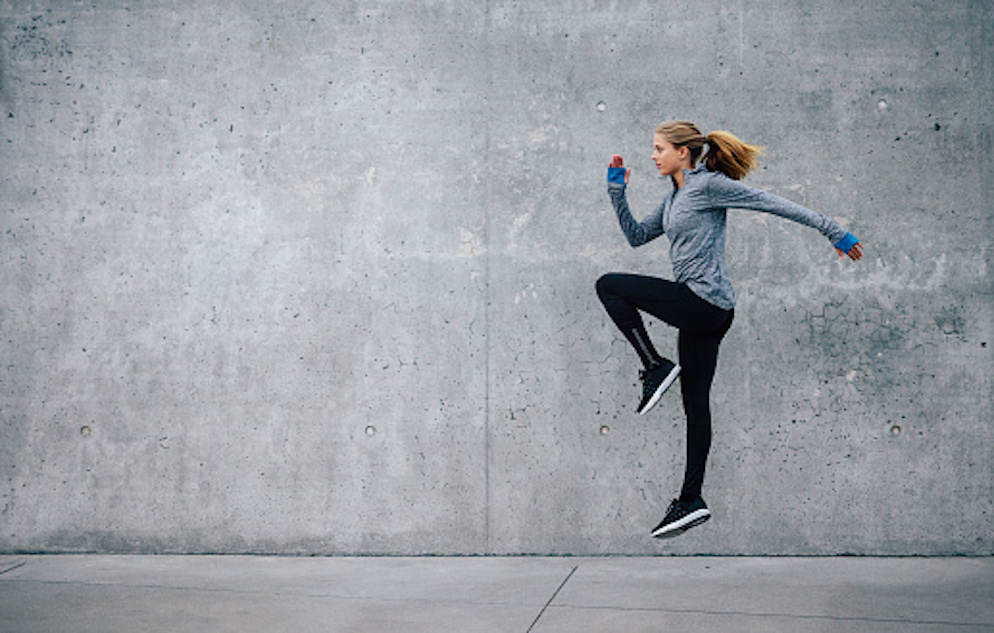 female jumping in a running pose in front of a concrete wall.