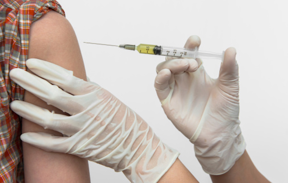 A syringe about to be injected into the upper arm of a patient by a person wearing white latex gloves.