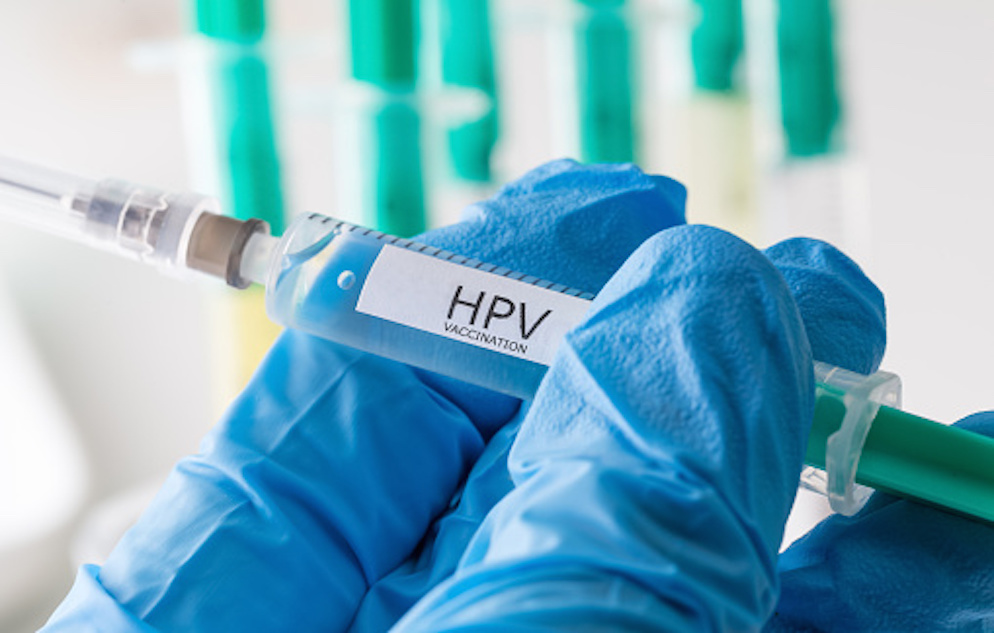 Fingers covered in blue gloves and holding a clear and teal syringe with a white label that reads: HPV VACCINATION