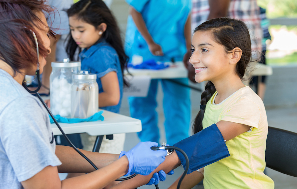 A smiling Hispanic, preteen girl having her blood pressure checked by a mid adult, African American woman