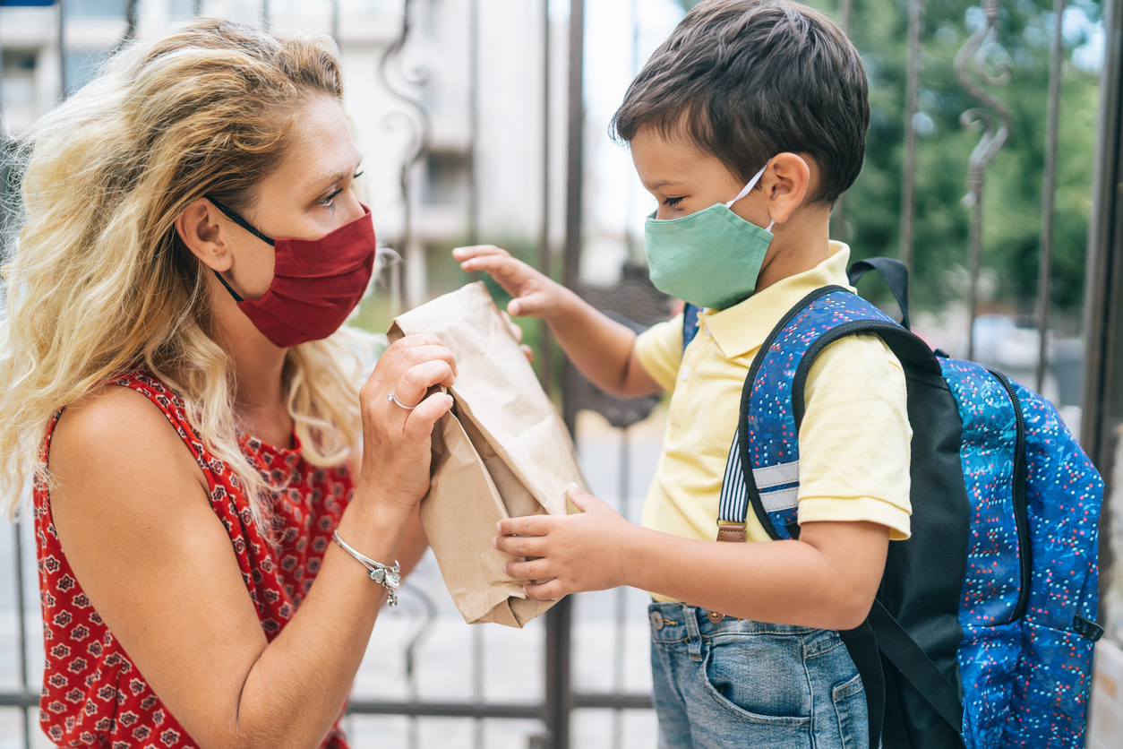 Mother gives sack lunch to son, both wearing protective face mask, during COVID-19