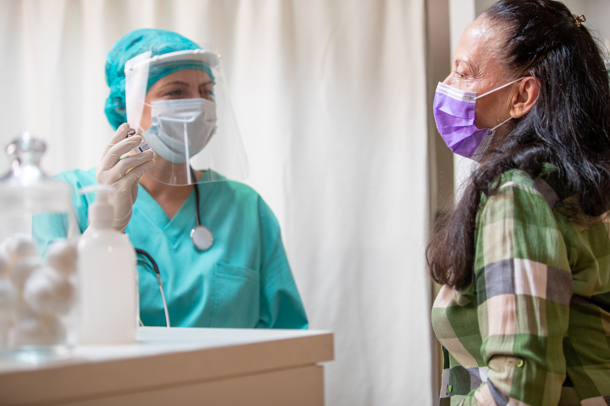 Senior woman wearing protective face mask sitting in doctor's office and waiting for vaccination, female doctor in protective workwear holding vial