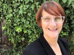 A white woman with red brown hair and clear glasses and a black suit jacket in front of an ivy wall, smiles.”