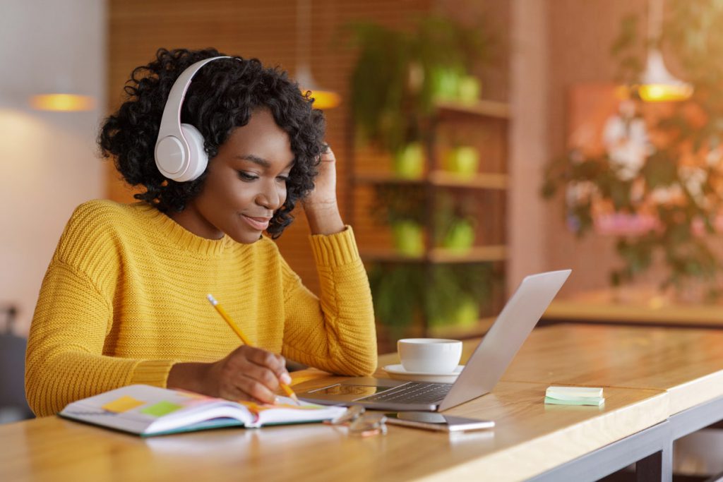 Student wearing yellow sweater with white headphones on, sitting in front of an open laptop and taking notes